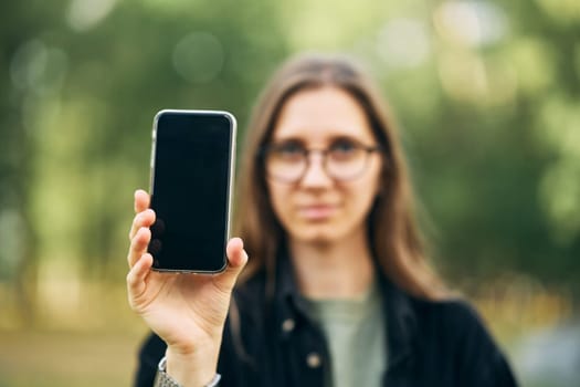 Young girl holds a phone in her hand showing it screen towards camera in the park. High quality photo
