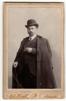 JIHLAVA, AUSTRIA-HUNGARY - CIRCA 1905: Vintage cabinet card shows dandy man wears bowler hat, luxury garment and holds walking stick and cigar. Antique black white photo.