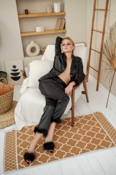 Woman glamorous pajamas. A beautiful blonde in black pajamas is sitting relaxed in a chair in black pajamas with feathers. Beige stones are all over the interior