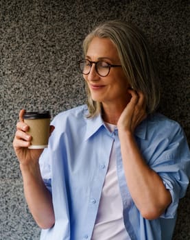 Joyful and stylish senior woman with coffee cup in her hand, standing near wall in urban city setting. She radiates happiness and relaxation, making it the perfect image to illustrate carefree and content lifestyle. High quality photo