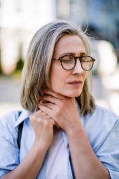 Sad senior woman wearing glasses holding her sore throat outdoors across the city, depicting the concept of illness and healthcare issues in aging populations.