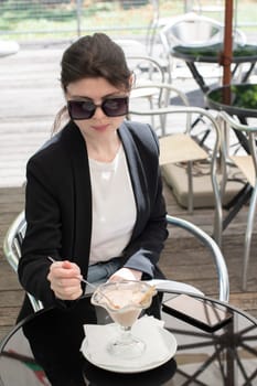 a business woman in sunglasses eats her favorite delicious ice cream, dressed in a black elegant jacket, her smartphone lies on the table, she enjoys a break from work, High quality photo