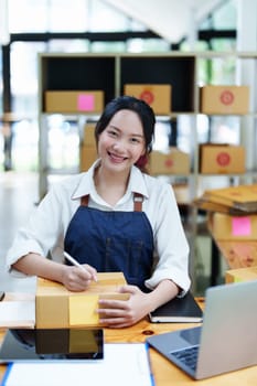 Starting small business entrepreneur of independent Asian woman smiling using computer laptop with cheerful success of online marketing package box items and SME delivery concept.