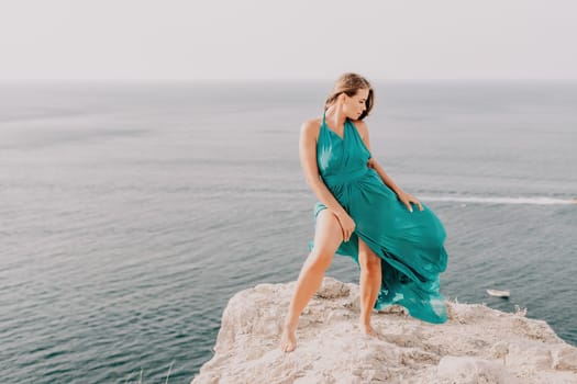 Side view a Young beautiful sensual woman in a mint long dress posing on a volcanic rock high above the sea during sunset. Girl on the nature on overcast sky background. Fashion photo