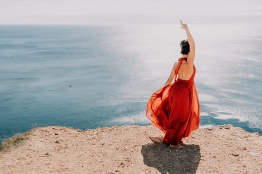 Side view a Young beautiful sensual woman in a red long dress posing on a rock high above the sea during sunrise. Girl on the nature on blue sky background. Fashion photo.