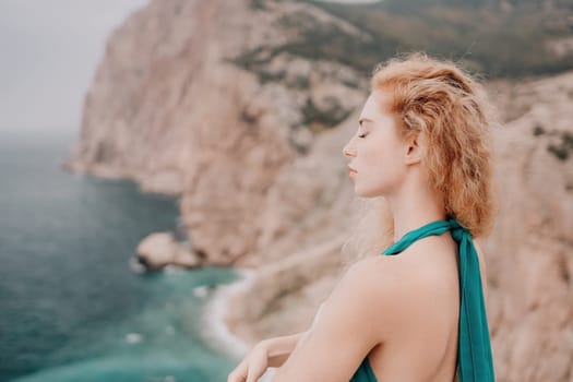 Side view a Young beautiful sensual woman in a mint long dress posing on a volcanic rock high above the sea during sunset. Girl on the nature on overcast sky background. Fashion photo