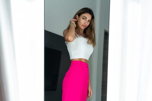 Stylish fit tanned beautiful woman in fashion pink pants and top posing in mirror at home bedroom, preparing for party, dressing up