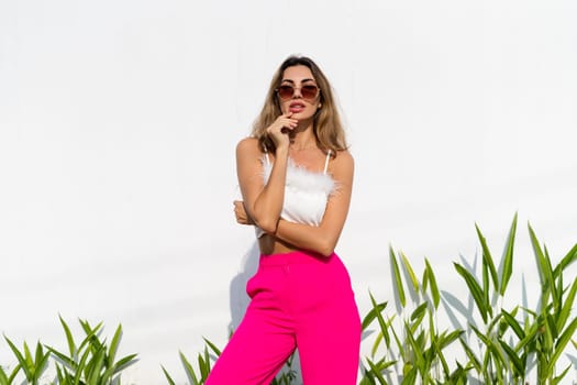 Stylish fit tanned beautiful woman in sunglasses, fashion pink pants and white top posing outdoor at white wall tropical leaves background, sunny day, natural light