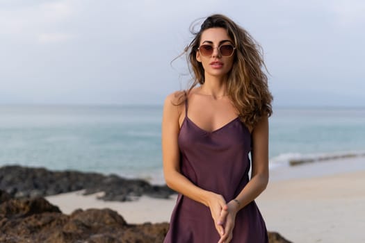 Stylish romantic tender sensual woman in silk dress and sunglasses on the beach at sunset