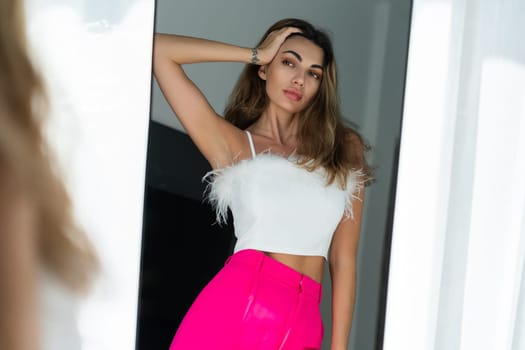 Stylish fit tanned beautiful woman in fashion pink pants and top posing in mirror at home bedroom, preparing for party, dressing up