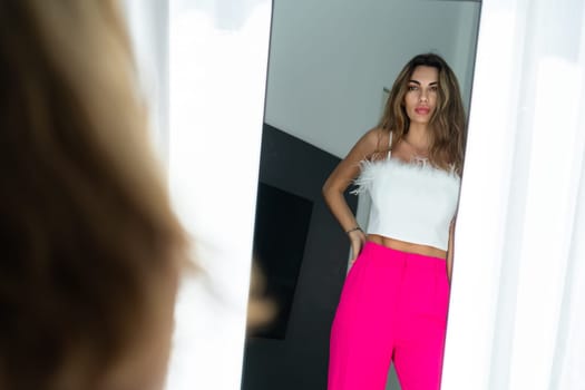 Stylish fit tanned beautiful woman in fashion pink pants and top posing in mirror at home bedroom, preparing for party, dressing up