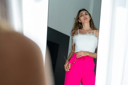 Stylish fit tanned beautiful woman in fashion pink pants and top posing in mirror at home bedroom, preparing for party, dressing up