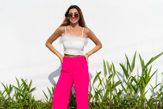 Stylish fit tanned beautiful woman in sunglasses, fashion pink pants and white top posing outdoor at white wall tropical leaves background, sunny day, natural light