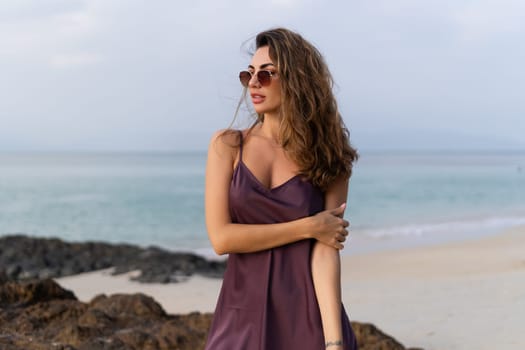 Stylish romantic tender sensual woman in silk dress and sunglasses on the beach at sunset