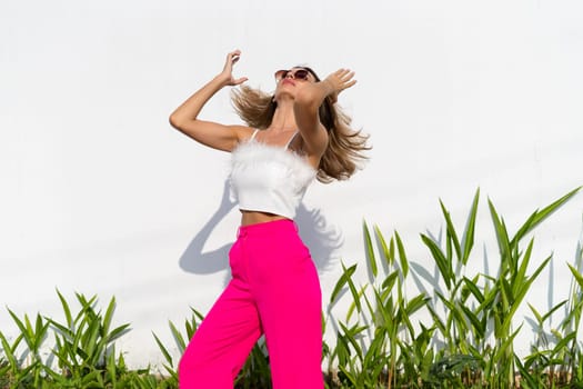 Stylish fit tanned beautiful woman in sunglasses, fashion pink pants and white top posing outdoor at white wall tropical leaves background, sunny day, natural light