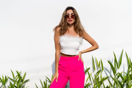Stylish fit tanned beautiful woman in sunglasses, fashion pink pants and white top posing outdoor at white wall tropical leaves background, sunny day, natural light