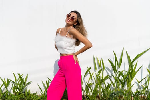 Stylish fit tanned beautiful woman in sunglasses, fashion pink pants and white top posing outdoor at white wall tropical leaves background, sunny day, natural light