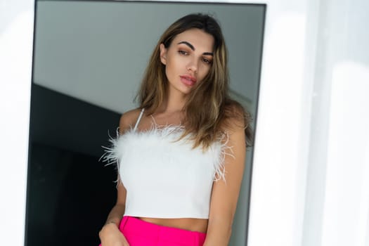 Stylish fit tanned beautiful woman in fashion pink pants and top posing in mirror at home bedroom, preparing for party, dressing up