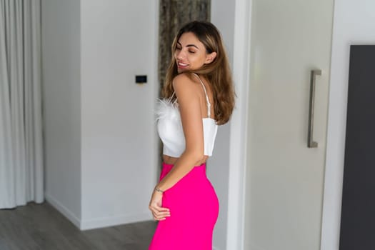 Stylish fit tanned beautiful woman in fashion pink pants and top posing in bedroom at home, preparing for party, dressing up