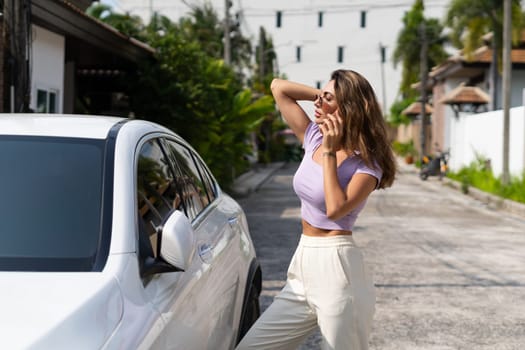 Successful smiling fit tanned attractive woman in casual wear is using her smart phone while standing near luxury modern car outdoors.