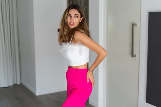 Stylish fit tanned beautiful woman in fashion pink pants and top posing in bedroom at home, preparing for party, dressing up