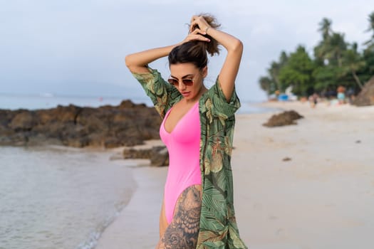 Stylish romantic tender athletic slim sensual woman in pink swimsuit, tropical kimono and sunglasses on the beach at sunset