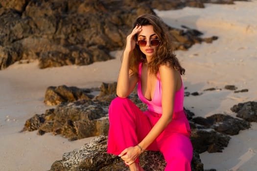 Fashion stylish beautiful sensual young woman, slim, in trendy bright pink pants and bodysuit on a tropical beach at sunset posing in sunglasses