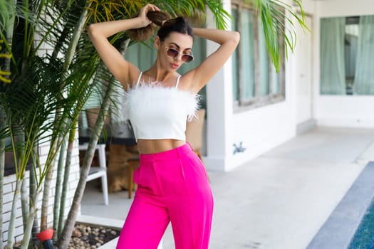 Stylish fit tanned beautiful woman in sunglasses, fashion pink pants and white top posing outdoor at luxury tropical villa