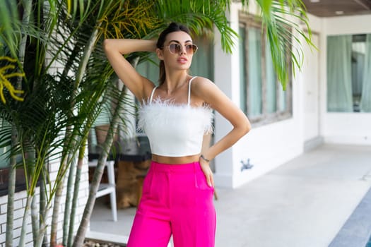 Stylish fit tanned beautiful woman in sunglasses, fashion pink pants and white top posing outdoor at luxury tropical villa