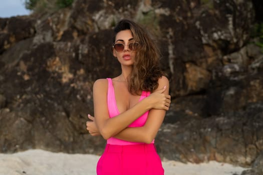 Fashion stylish beautiful sensual young woman, slim, in trendy bright pink pants and bodysuit on a tropical beach at sunset posing in sunglasses