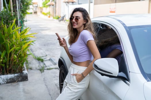 Successful smiling fit tanned attractive woman in casual wear is using her smart phone while standing near luxury modern car outdoors.