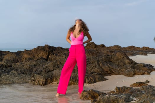 Fashion stylish beautiful sensual young woman, slim, in trendy bright pink pants and bodysuit on a tropical beach at sunset posing in sunglasses