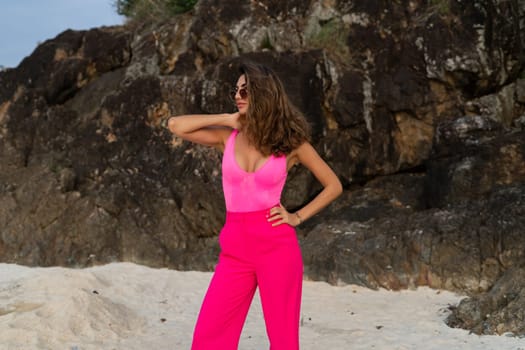 Fashion stylish beautiful sensual young woman, slim, in trendy bright pink pants and bodysuit on a tropical beach at sunset posing in sunglasses