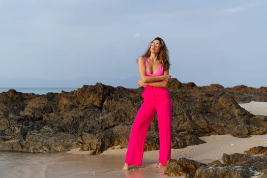 Fashion stylish beautiful sensual young woman, slim, in trendy bright pink pants and bodysuit on a tropical beach at sunset posing in sunglasses