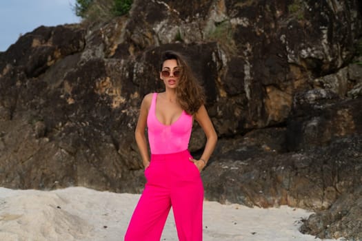 Fashion stylish beautiful sensual young woman, slim, in trendy bright pink pants and bodysuit on a tropical beach at sunset posing in sunglasses