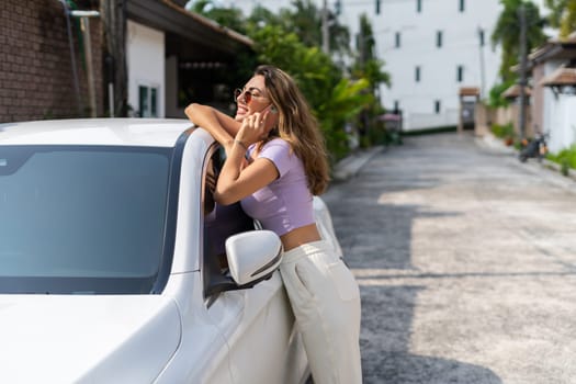 Successful smiling fit tanned attractive woman in casual wear is using her smart phone while standing near luxury modern car outdoors.