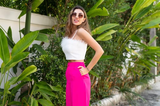 Stylish fit tanned beautiful woman in sunglasses, fashion pink pants and white top posing outdoor at luxury tropical villa