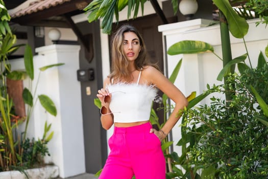 Stylish fit tanned beautiful woman in sunglasses, fashion pink pants and white top posing outdoor at luxury tropical villa