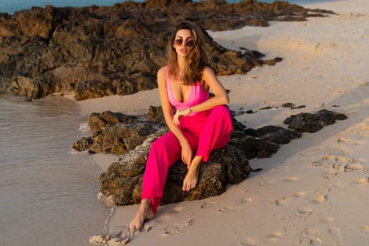 Fashion stylish beautiful sensual young woman, slim, in trendy bright pink pants and bodysuit on a tropical beach at sunset posing in sunglasses