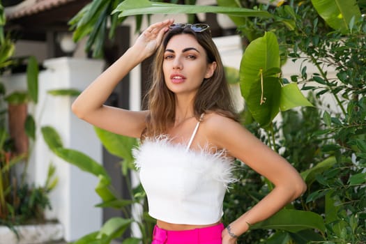 Stylish fit tanned beautiful woman in sunglasses, fashion pink pants and white top posing outdoor at luxury tropical villa