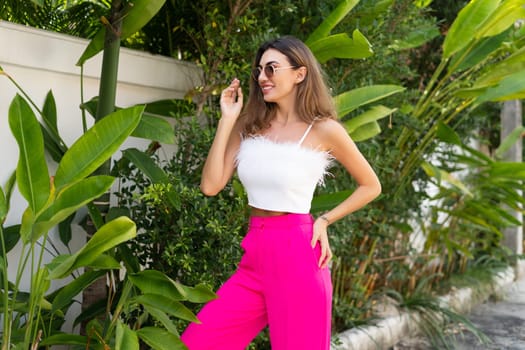 Stylish fit tanned beautiful woman in sunglasses, fashion pink pants and white top posing outdoor at luxury tropical villa