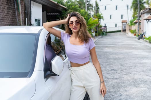 Successful smiling fit tanned attractive woman in casual wear is using her smart phone while standing near luxury modern car outdoors.