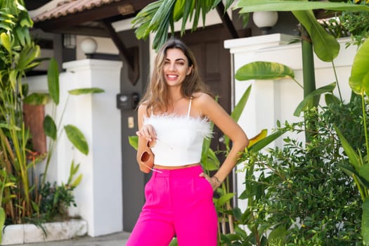 Stylish fit tanned beautiful woman in sunglasses, fashion pink pants and white top posing outdoor at luxury tropical villa