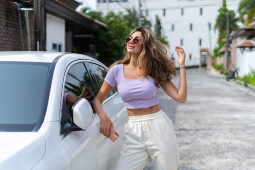 Successful smiling fit tanned attractive woman in casual wear is using her smart phone while standing near luxury modern car outdoors.