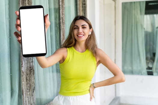 Portrait of cheerful woman standing outdoor with smartphone and headphones holding phone with white empty big screen with happy smile