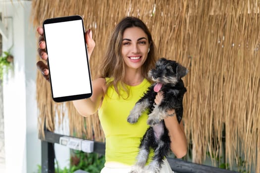 Portrait of cheerful woman standing outdoor with smartphone and headphones, with miniature schnauzer puppy dog, holding phone with white empty big screen with happy smile