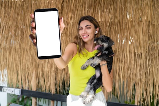 Portrait of cheerful woman standing outdoor with smartphone and headphones, with miniature schnauzer puppy dog, holding phone with white empty big screen with happy smile