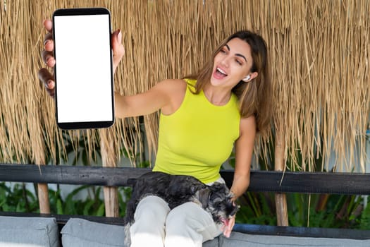 Portrait of cheerful woman standing outdoor with smartphone and headphones, with miniature schnauzer puppy dog, holding phone with white empty big screen with happy smile