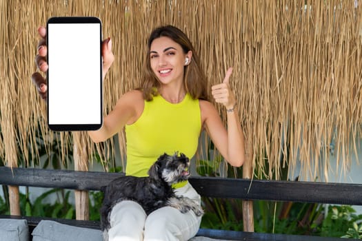 Portrait of cheerful woman standing outdoor with smartphone and headphones, with miniature schnauzer puppy dog, holding phone with white empty big screen with happy smile