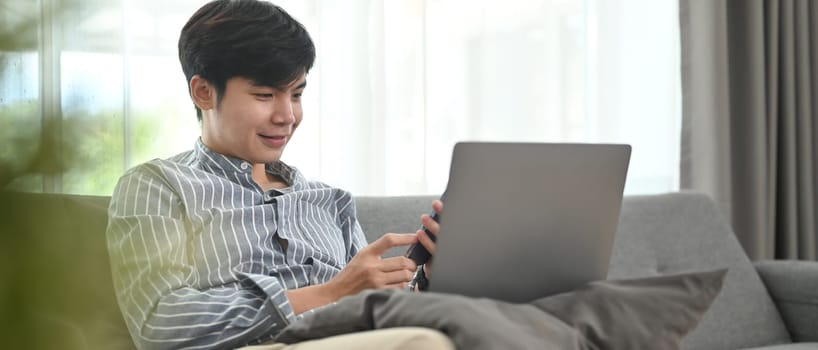 Happy millennial man relaxing on couch and enjoy browsing internet on laptop. People, leisure and technology concept.